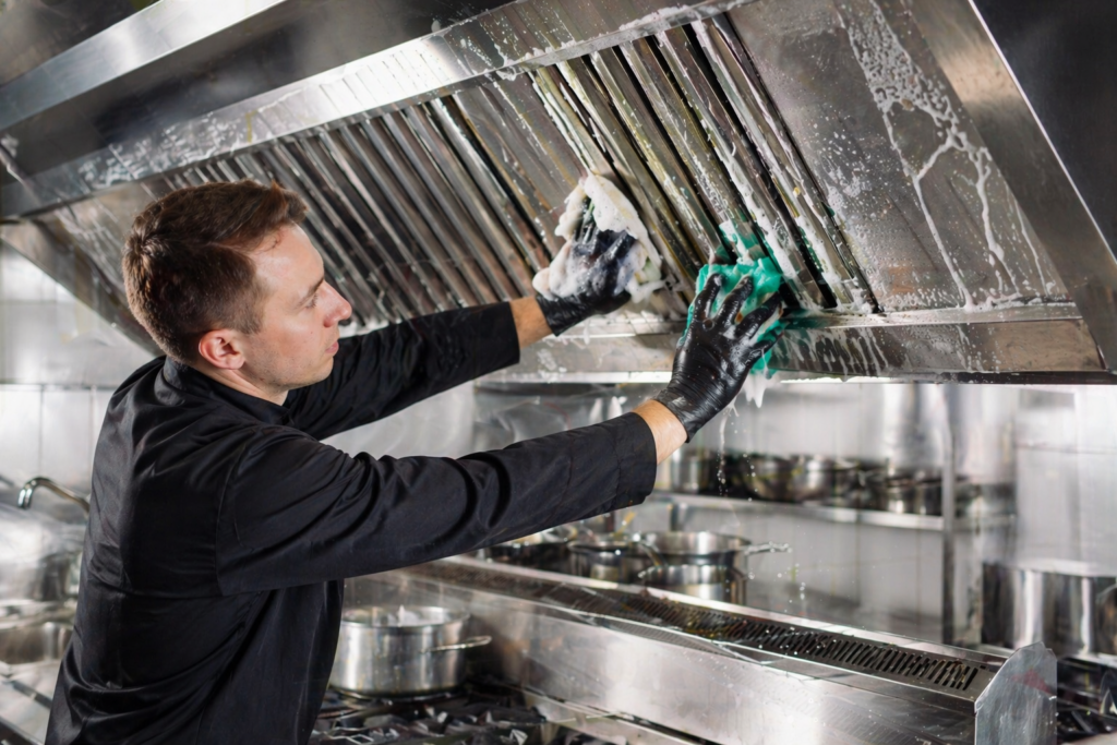 Technician performing kitchen duct cleaning in Dubai by scrubbing a commercial kitchen exhaust hood and grease filters with foam and cleaning tools.